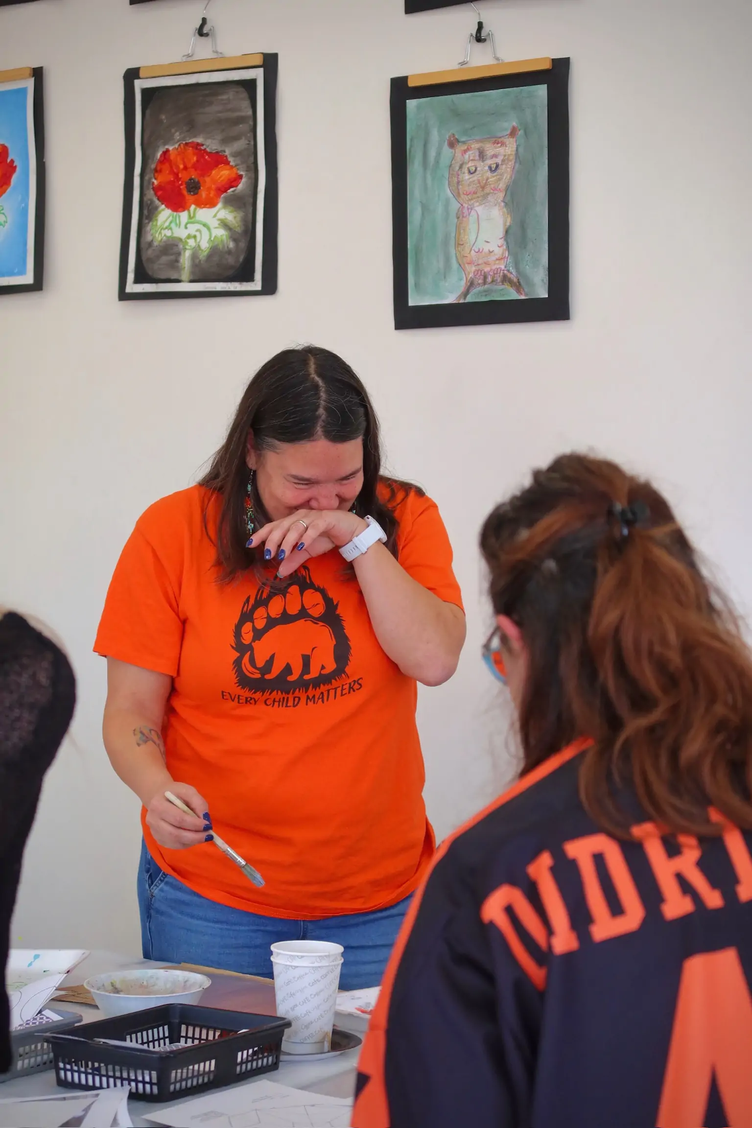 participant in an orange “Every Child Matters” shirt smiling and wiping their eyes while working on an art activity, with another attendee seated in front of them and student artwork hanging on the wall