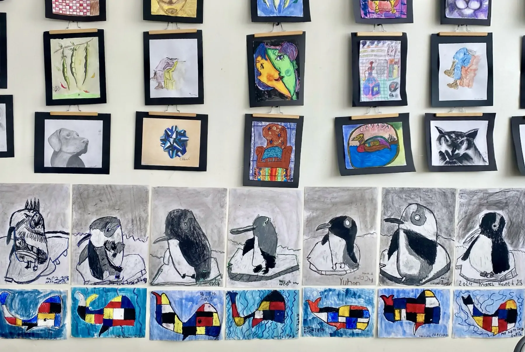 Wall display of children’s artwork at Aliki’s Art House featuring penguin drawings, abstract whales, and mixed-media fine art pieces.