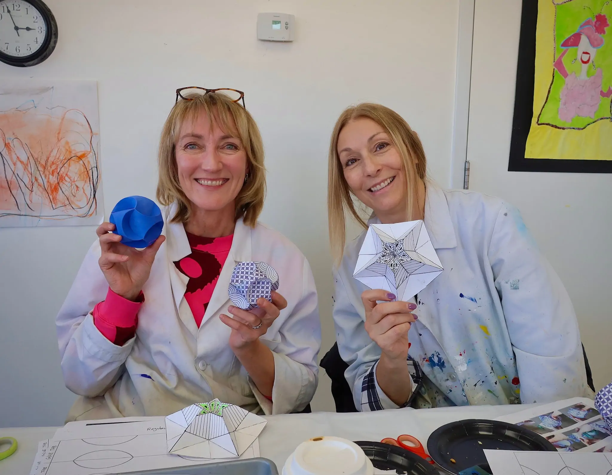 two adult students smiling and holding geometric paper art projects during an art class at Aliki’s Art House in Calgary