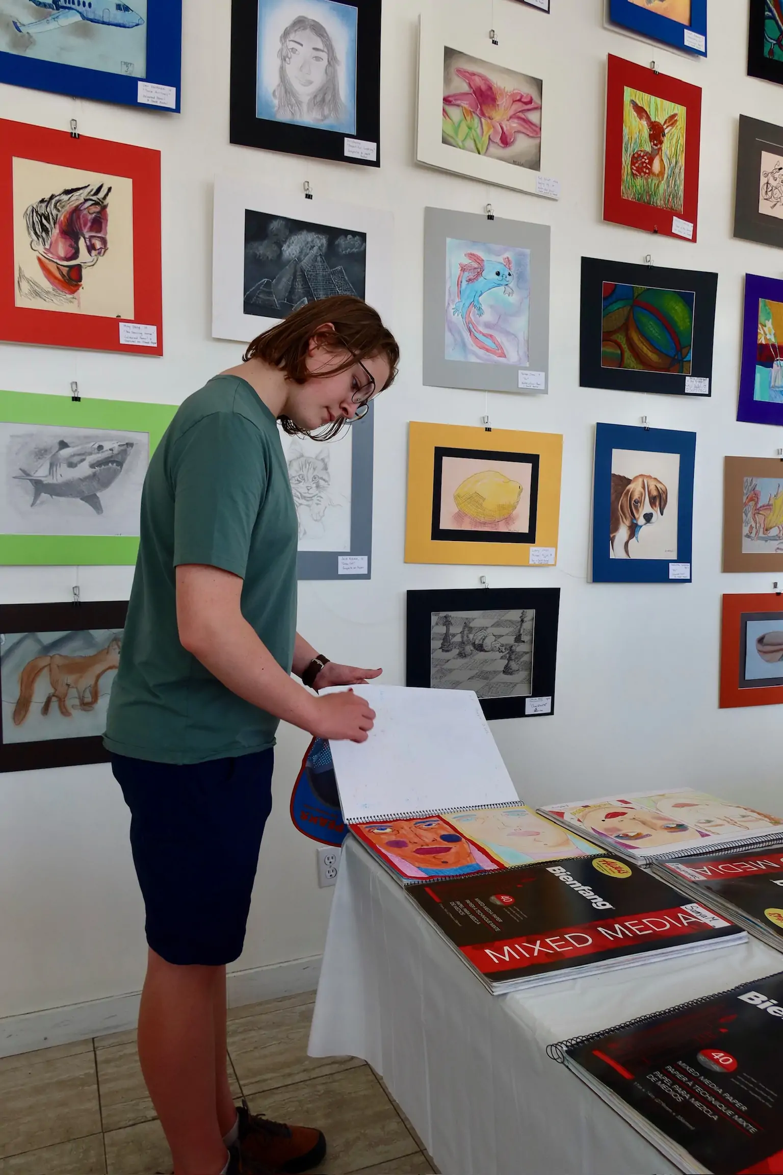 A teen student looks through a portfolio of artwork while standing beside a display wall filled with framed student pieces at Aliki’s Art House.