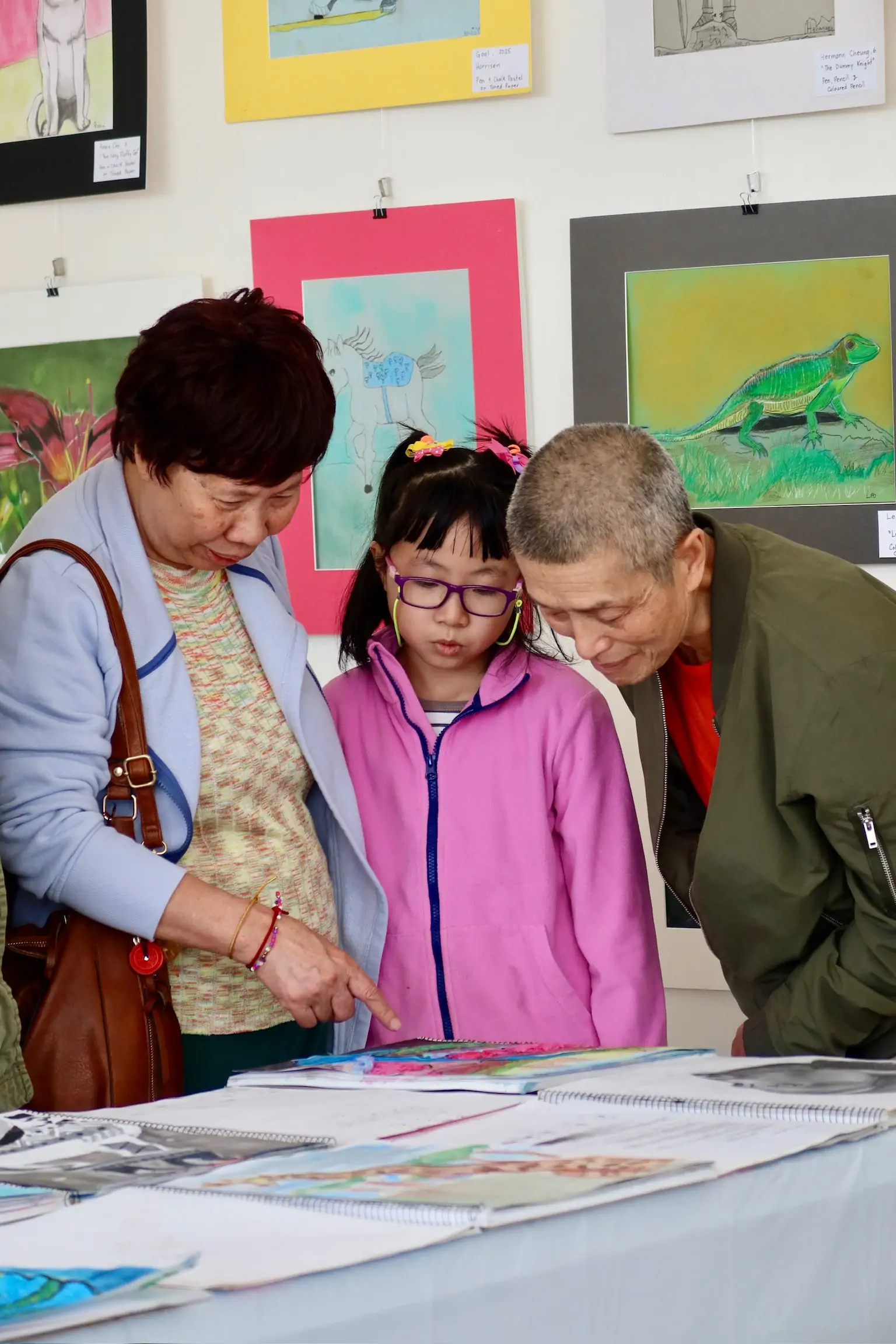 A child in a pink jacket looks at their artwork on the table with two adults during the student art showcase at Aliki’s Art House.