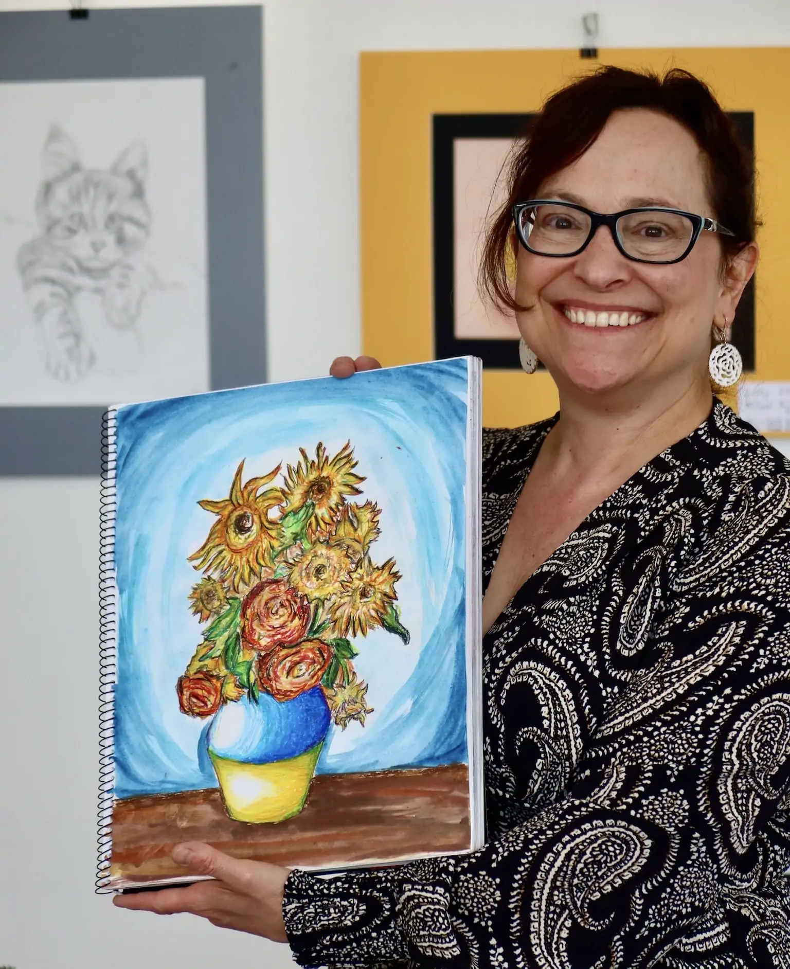 Diana, CEO of Aliki’s Art House, smiling and holding a student art piece featuring a colorful vase of sunflowers.