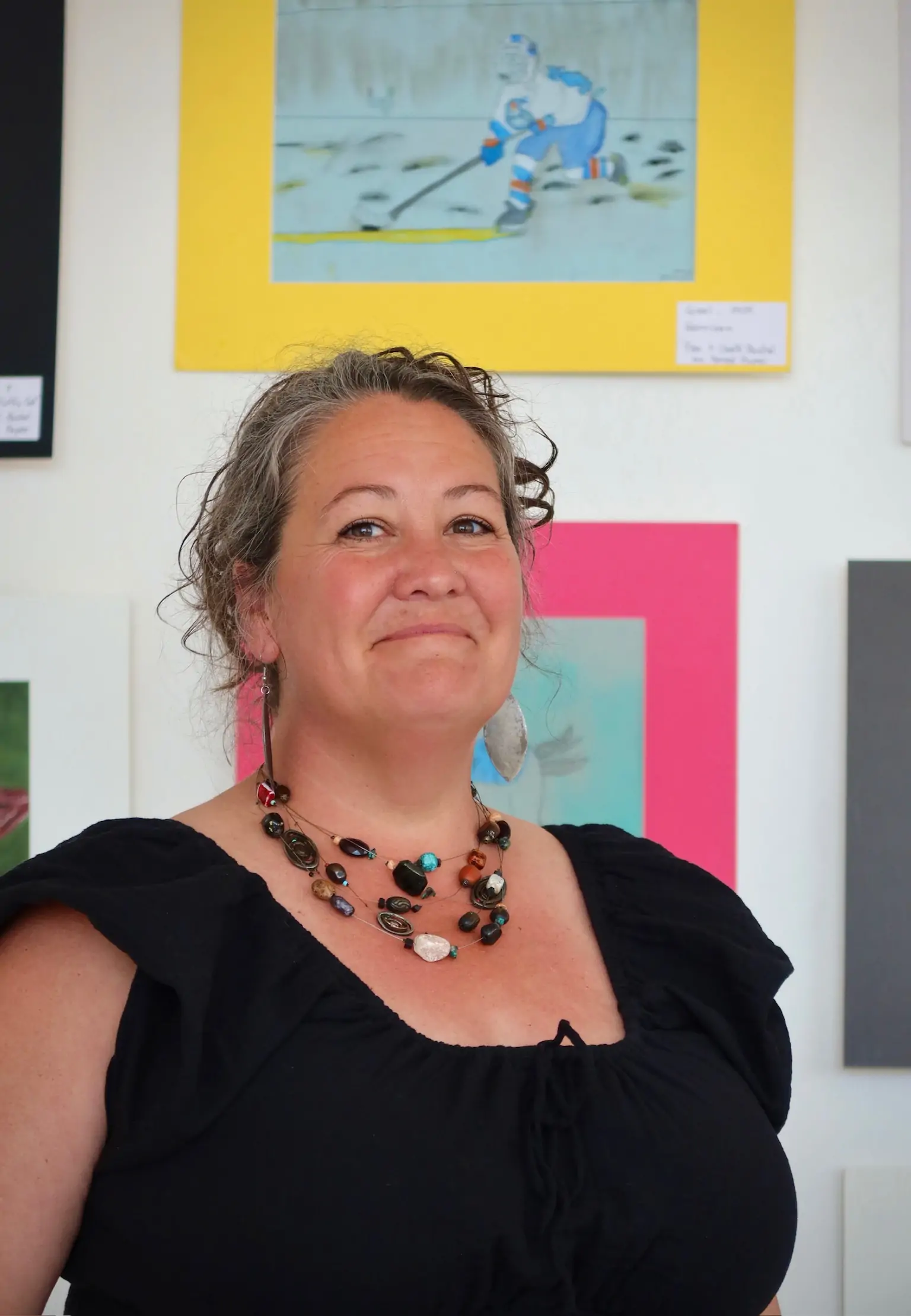 Alesha Buczny Brown, Bachelor of Fine Arts graduate specializing in jewelry and metals, smiling in front of colorful student artwork at Aliki’s Art House in Calgary