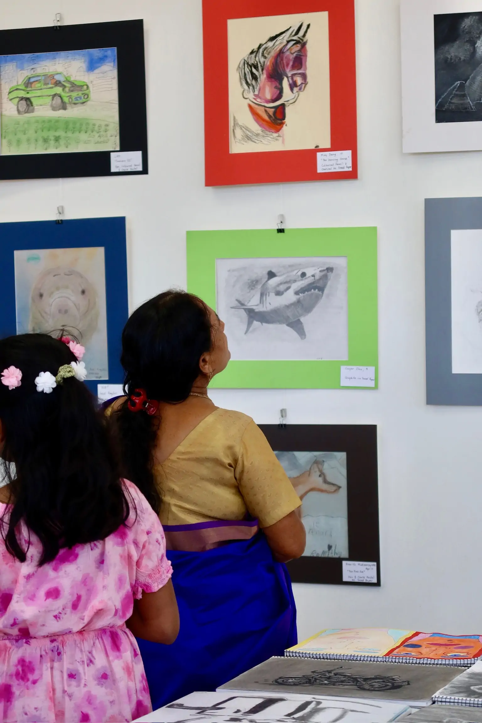 A child and an adult view framed student artwork displayed on the wall at Aliki’s Art House.