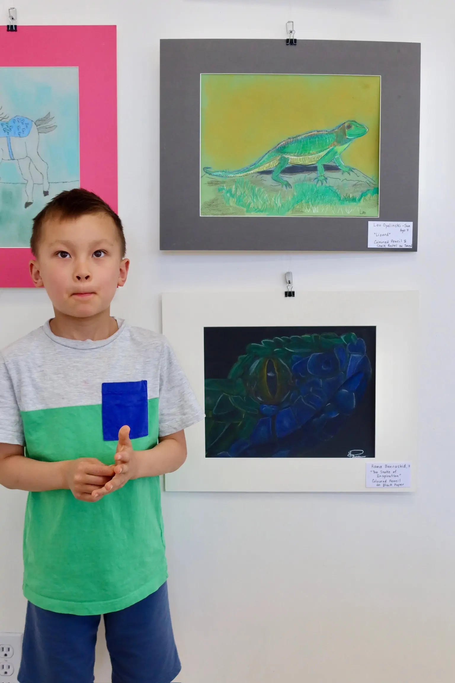 A young student stands beside their artwork displayed on the wall at Aliki’s Art House.