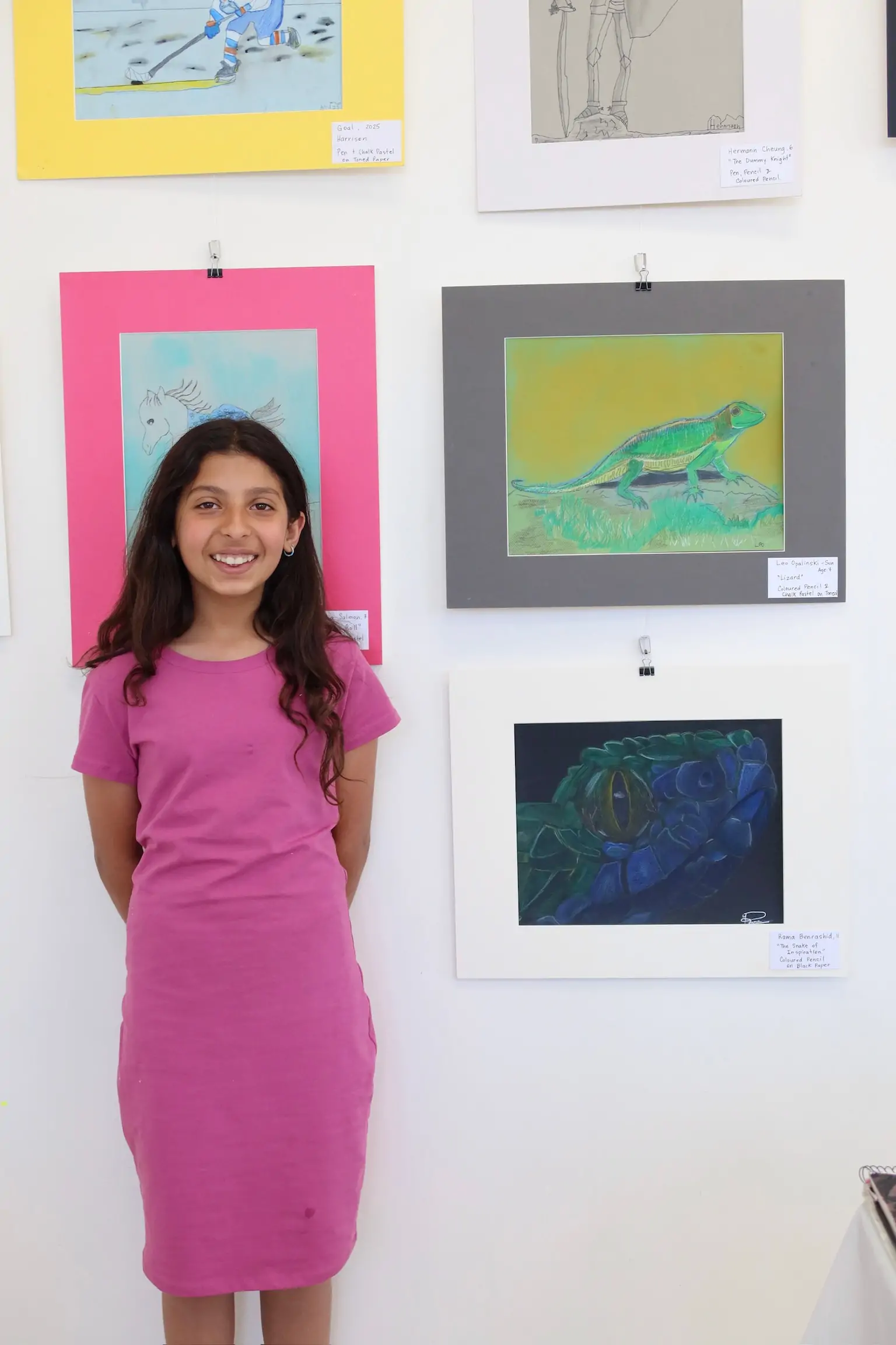 Smiling girl in a pink dress standing beside her framed artwork at the Aliki’s Art House student showcase.