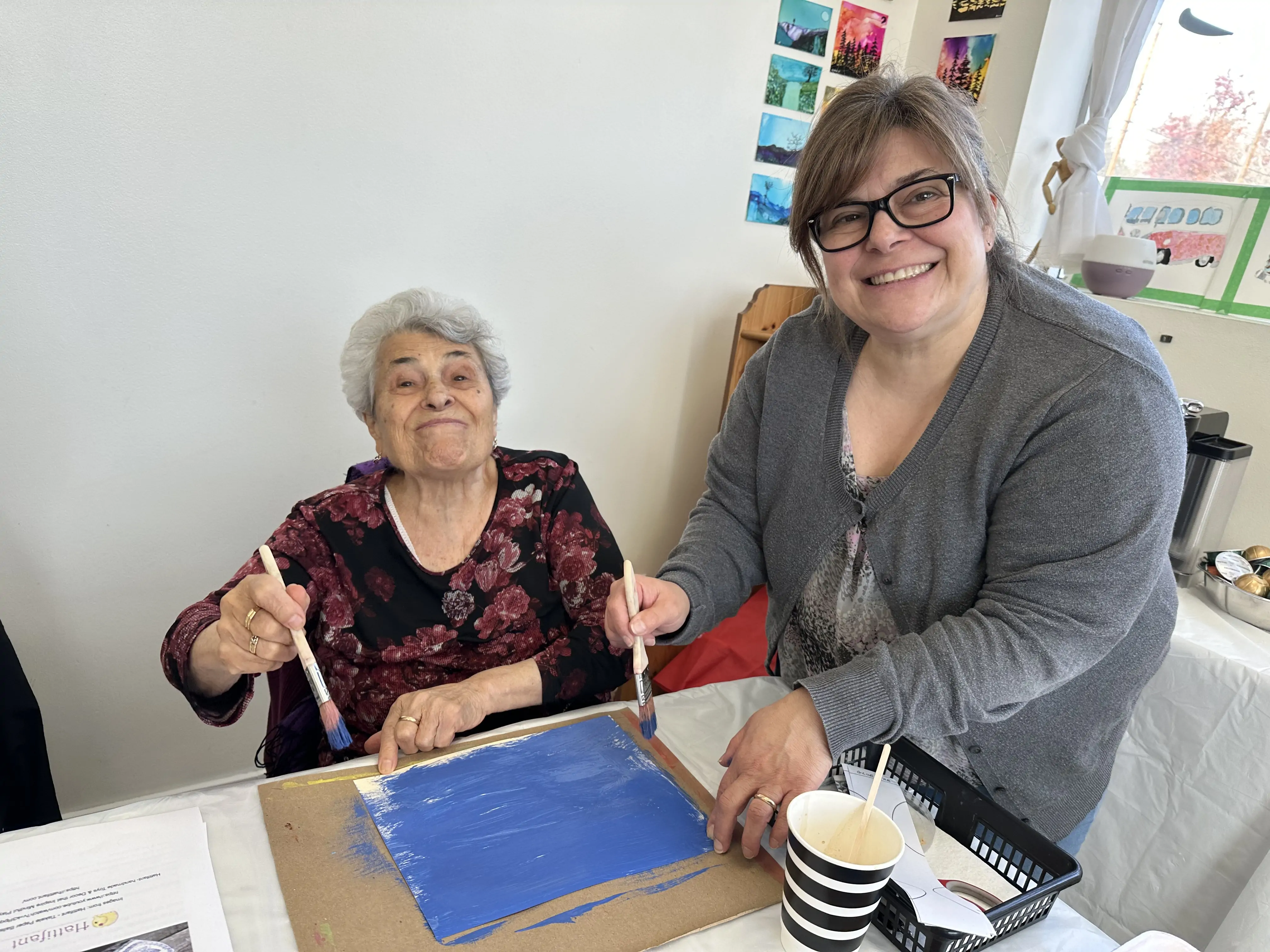 two women smiling while painting a blue background together during an art activity at Aliki’s Art House