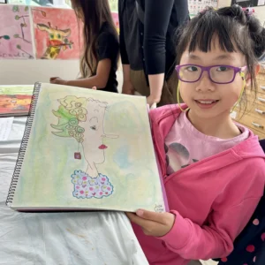 Young student smiling and holding her sketchbook drawing at Aliki’s Art House.